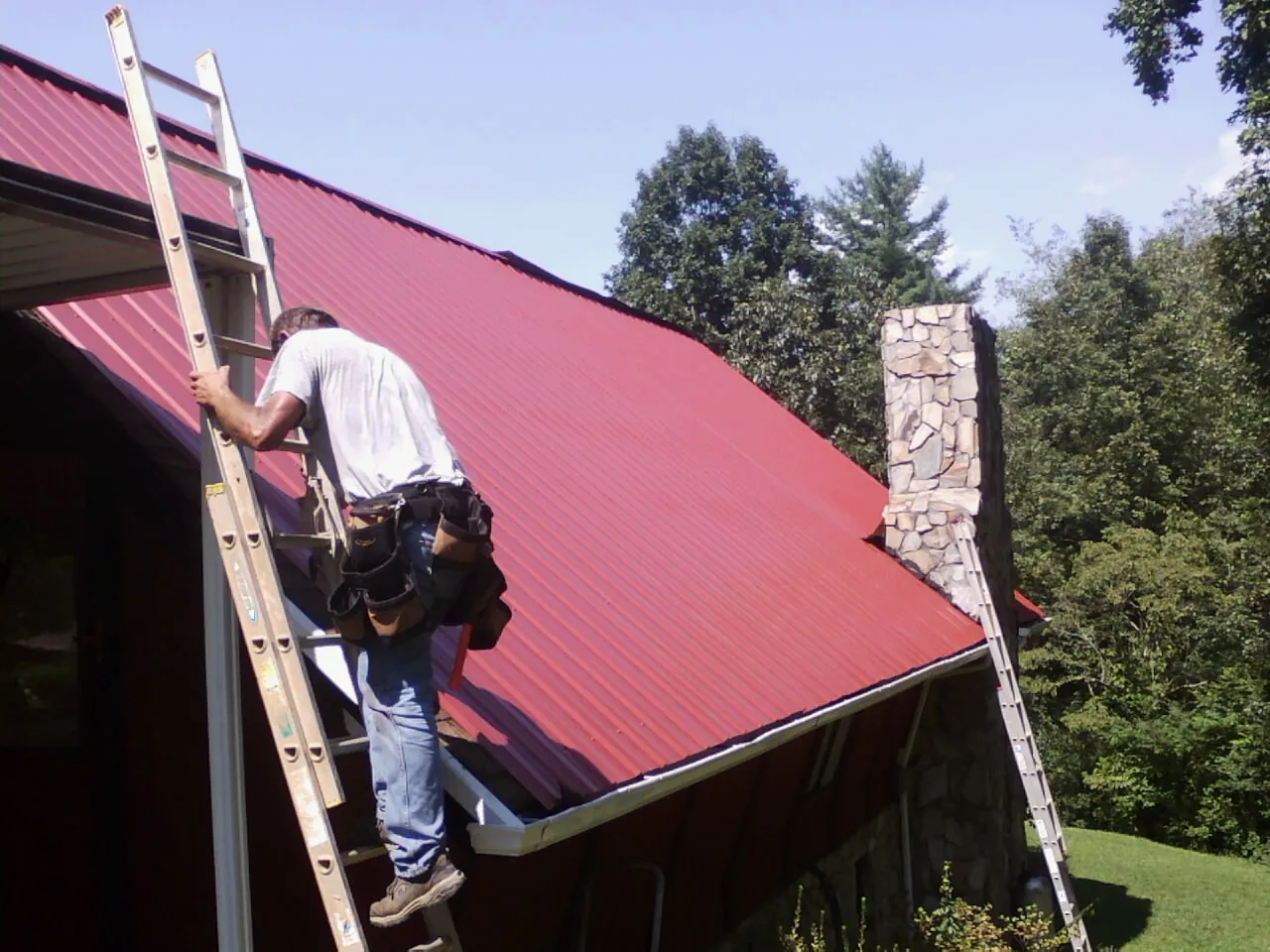Roofer on a ladder beside a red metal roof and stone chimney — Soffit & Fascia Repair by Roofing Brothers in Shaftsbury